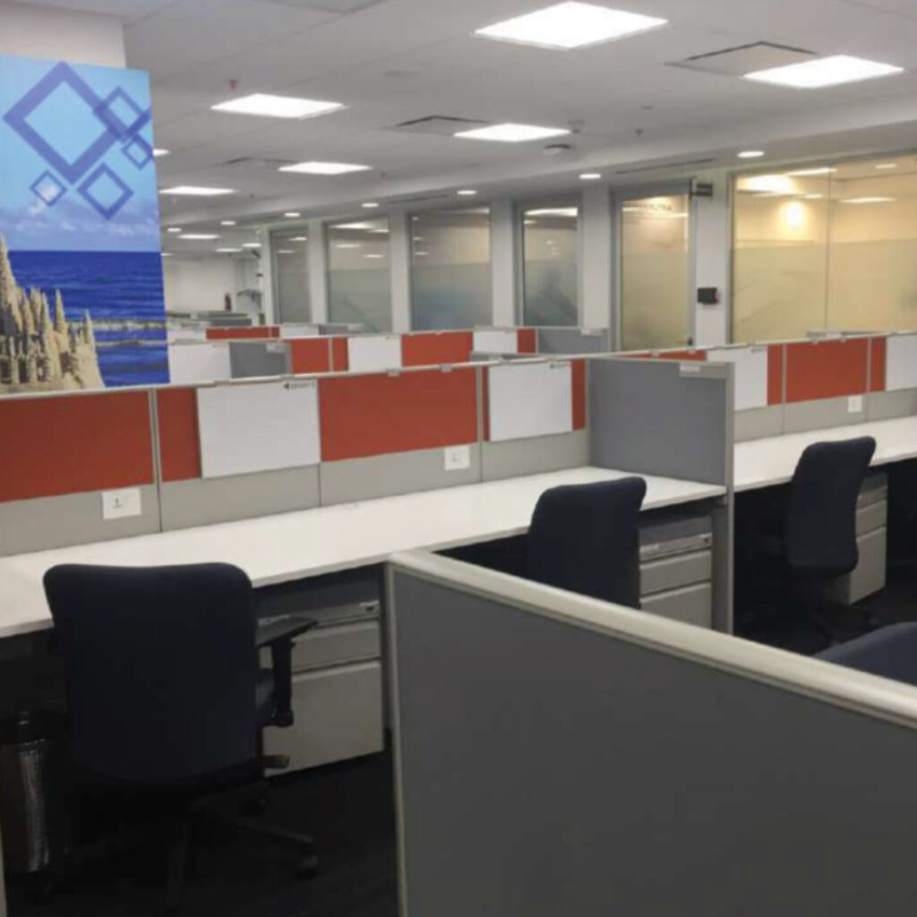 Open-plan office with cubicles and red partitions, row of desks and black office chairs under bright ceiling lights.