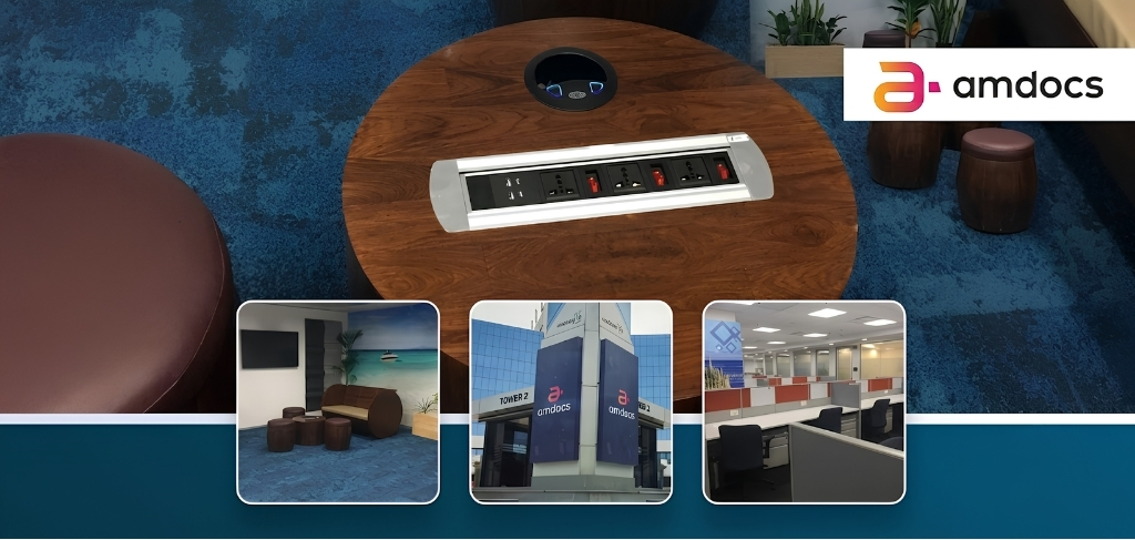 Round wooden table with built-in power outlets on a blue carpet, with a seating area and Amdocs branding in the corner.