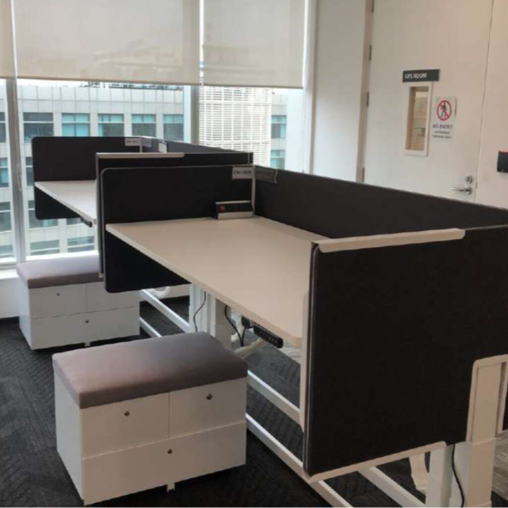 Open-plan office with white desks and dark cubicle partitions, large windows with blinds, and a city building visible outside the windows.